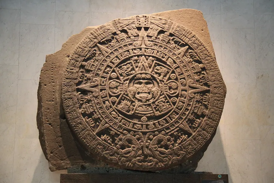 Massive circular basalt monolith carved with a central face surrounded by concentric glyph bands.