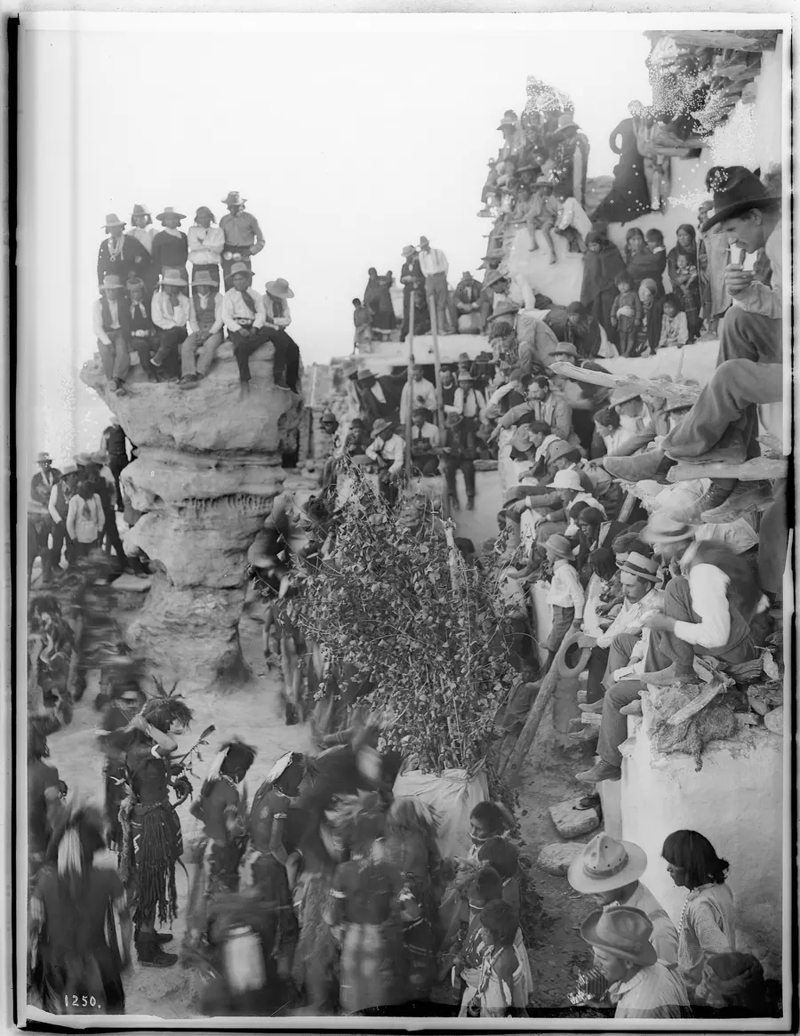 Crowd of Anglo spectators seated on pueblo rooftops watching a ceremony below.